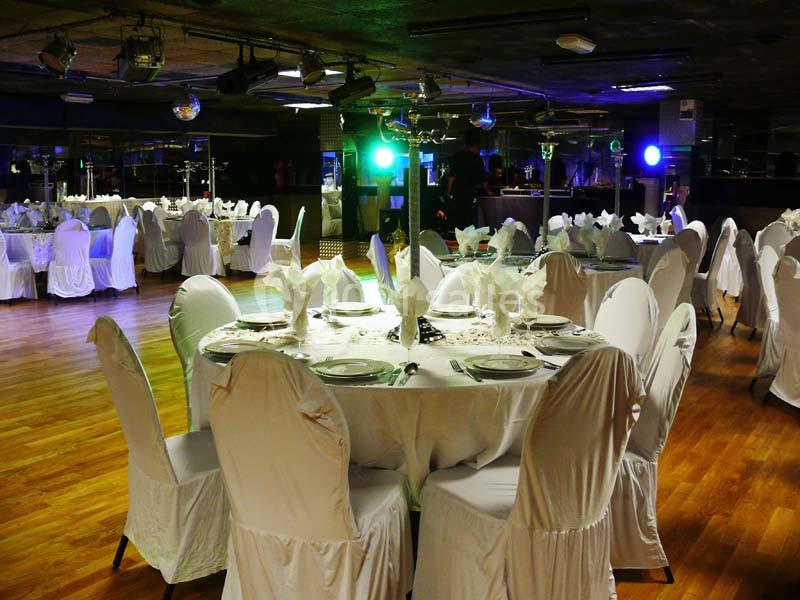 Salle de réception avec tables rondes dressées, nappes blanches et chaises couvertes, éclairage tamisé.