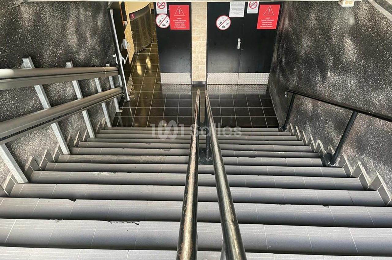 Escalier intérieur en carrelage gris avec rampes noires, menant à des portes fermées et des panneaux d'avertissement.