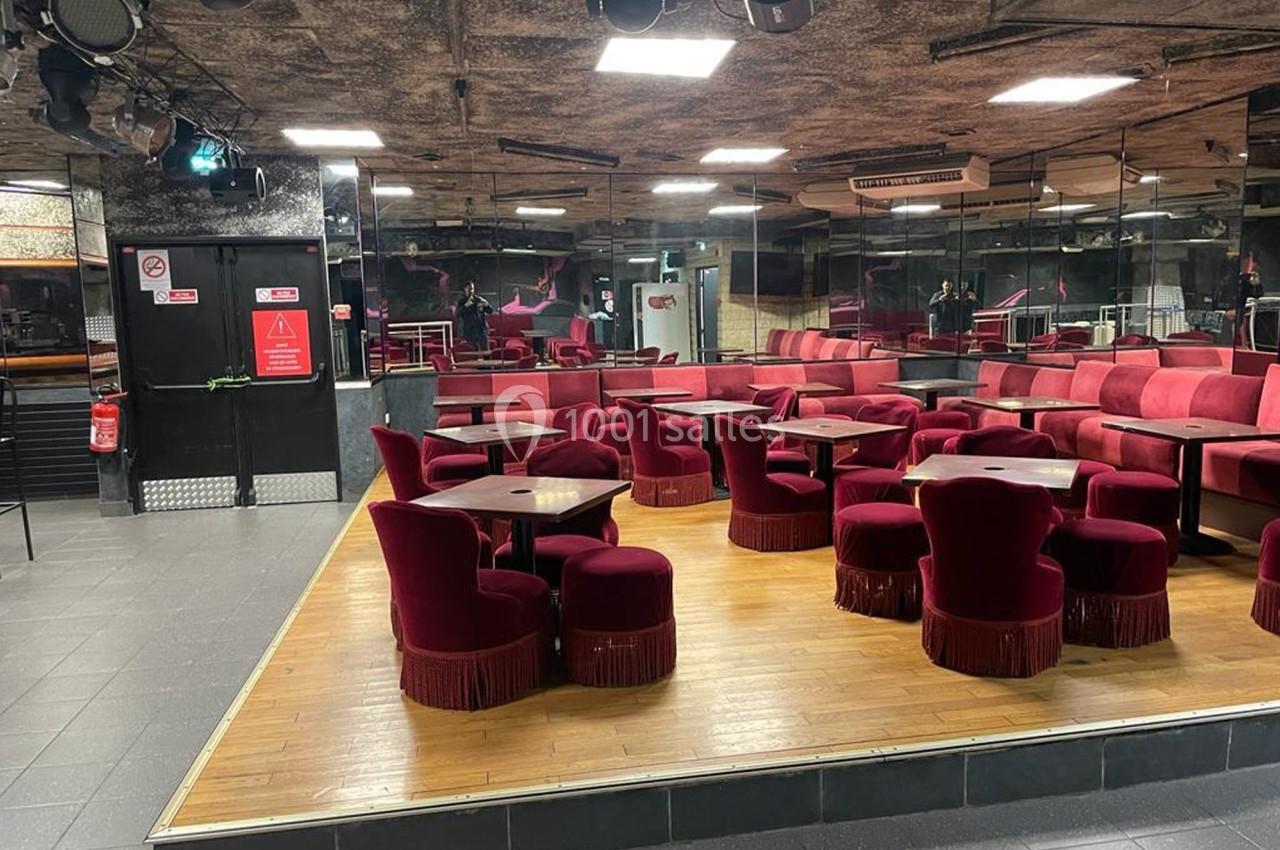 Salle avec des fauteuils rouges, tables en bois, sol partiellement carrelé et parquet, murs avec miroirs et éclairage tamisé.