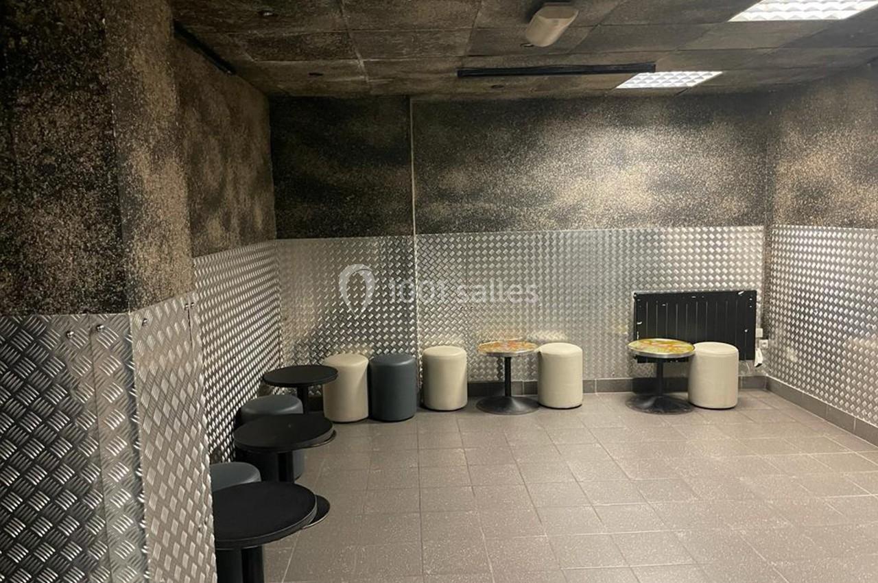 Salle avec murs texturés, sol carrelé, tables noires rondes et tabourets rembourrés disposés contre les murs.