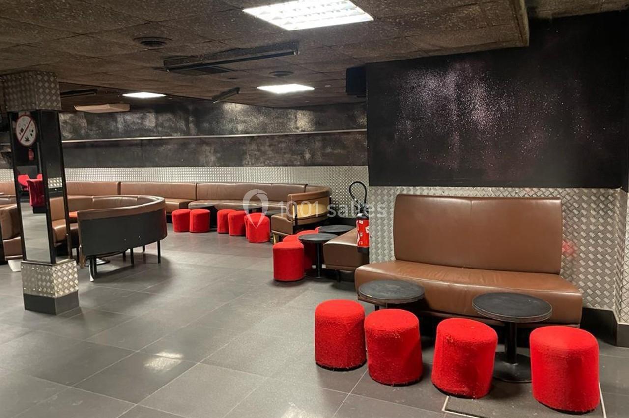 Salle avec banquettes marron, poufs rouges, tables basses noires et murs sombres, éclairée par des néons au plafond.
