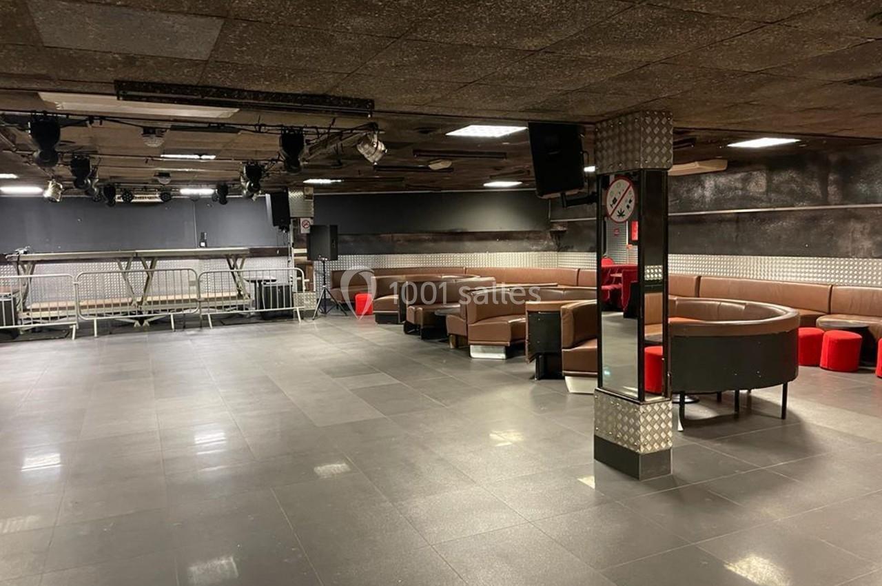 Salle vide avec sol en carrelage sombre, banquettes marron et rouges, éclairage tamisé et scène en arrière-plan.