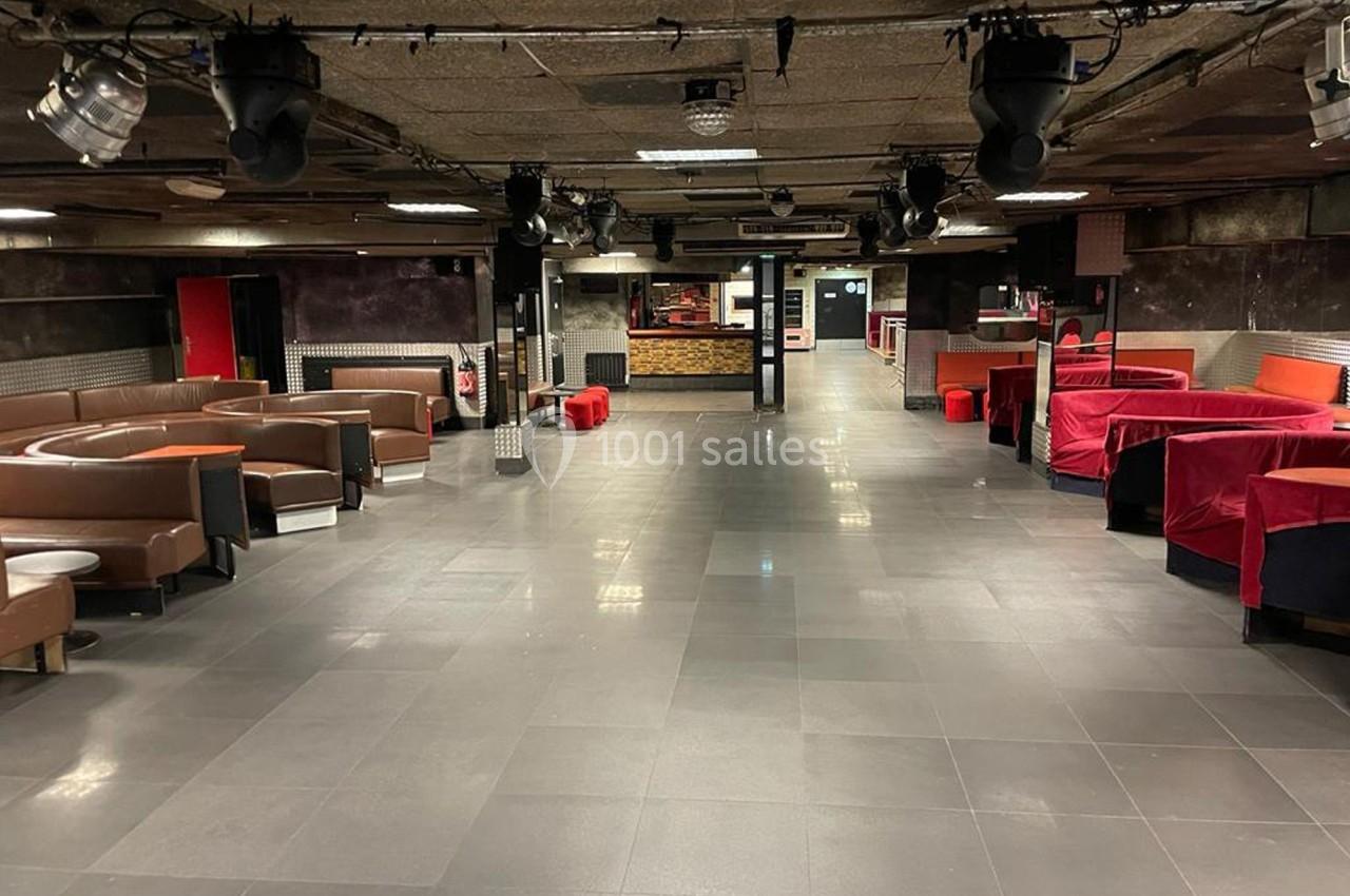 Salle vide avec canapés en cuir, tables basses et banquettes rouges, éclairage tamisé et sol en carrelage gris.