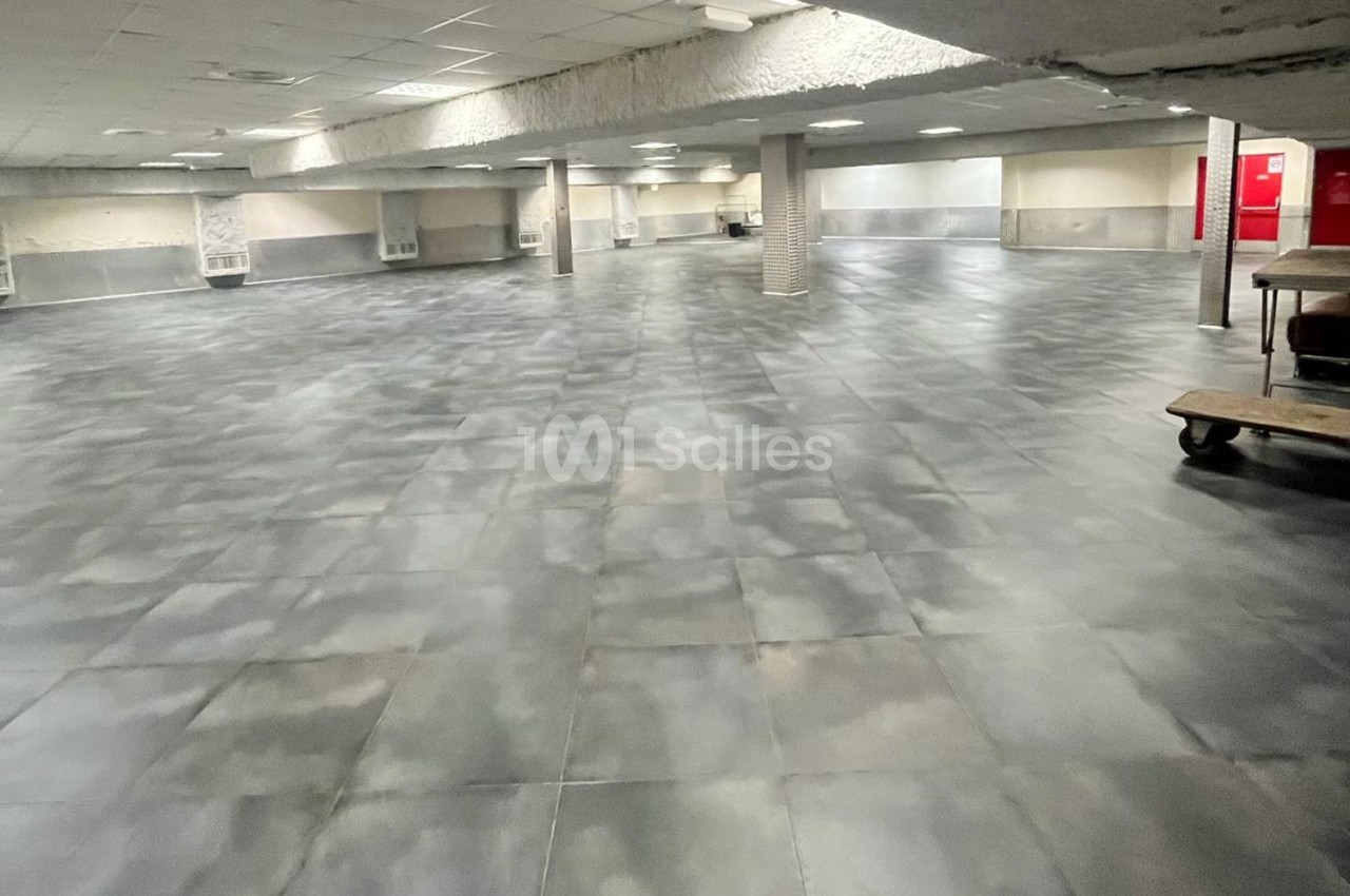 Salle vide avec un sol en carrelage gris, des murs clairs, un plafond bas et des colonnes réparties dans l'espace.
