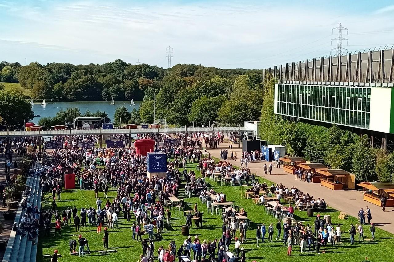 Location salle Nantes (Loire-Atlantique) - Exponantes Le Parc #19 Foule rassemblée dans un espace vert avec des stands, près d'un bâtiment moderne et d'un lac bordé d'arbres.
