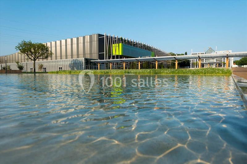 Location salle Nantes (Loire-Atlantique) - Exponantes Le Parc #6 Vue d'un bâtiment moderne avec une façade vitrée, entouré d'un plan d'eau et de végétation sous un ciel dégagé.