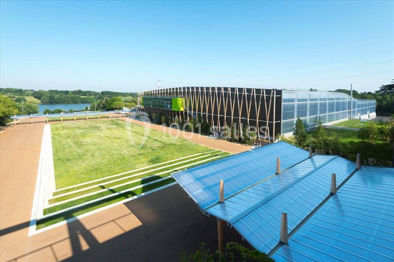 Location salle Nantes (Loire-Atlantique) - Exponantes Le Parc #7 Vue d'un bâtiment moderne entouré de verdure, avec un toit en verre au premier plan et un lac en arrière-plan.