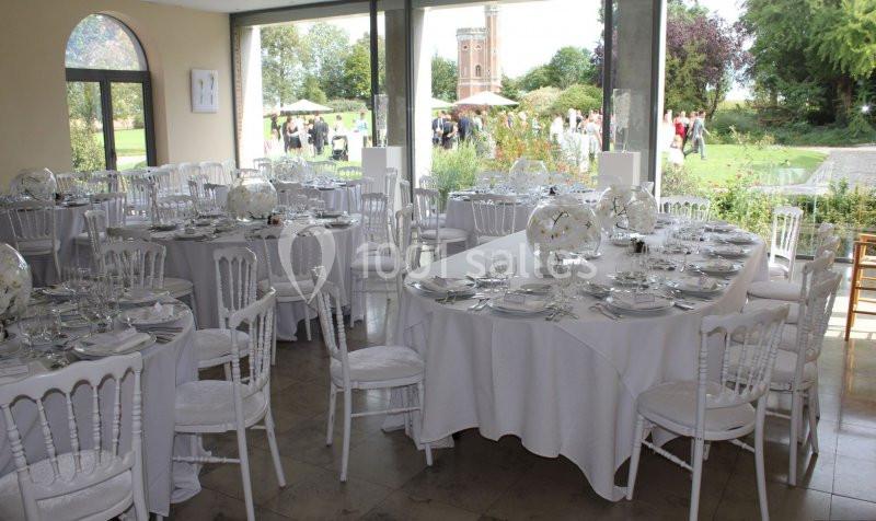 Location salle Donceel (Liège) - Château de Limont #3 Salle de réception lumineuse avec tables rondes et ovales dressées, vue sur un jardin et des invités à l'extérieur.