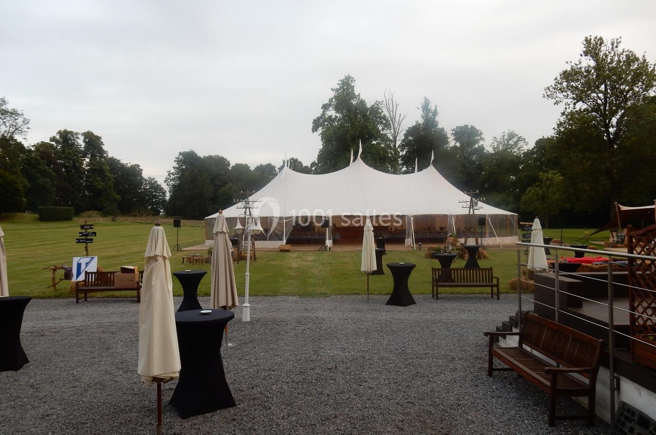 Location salle Donceel (Liège) - Château de Limont #16 Grande tente blanche installée sur une pelouse entourée d'arbres, avec des tables et parasols disposés à l'extérieur.