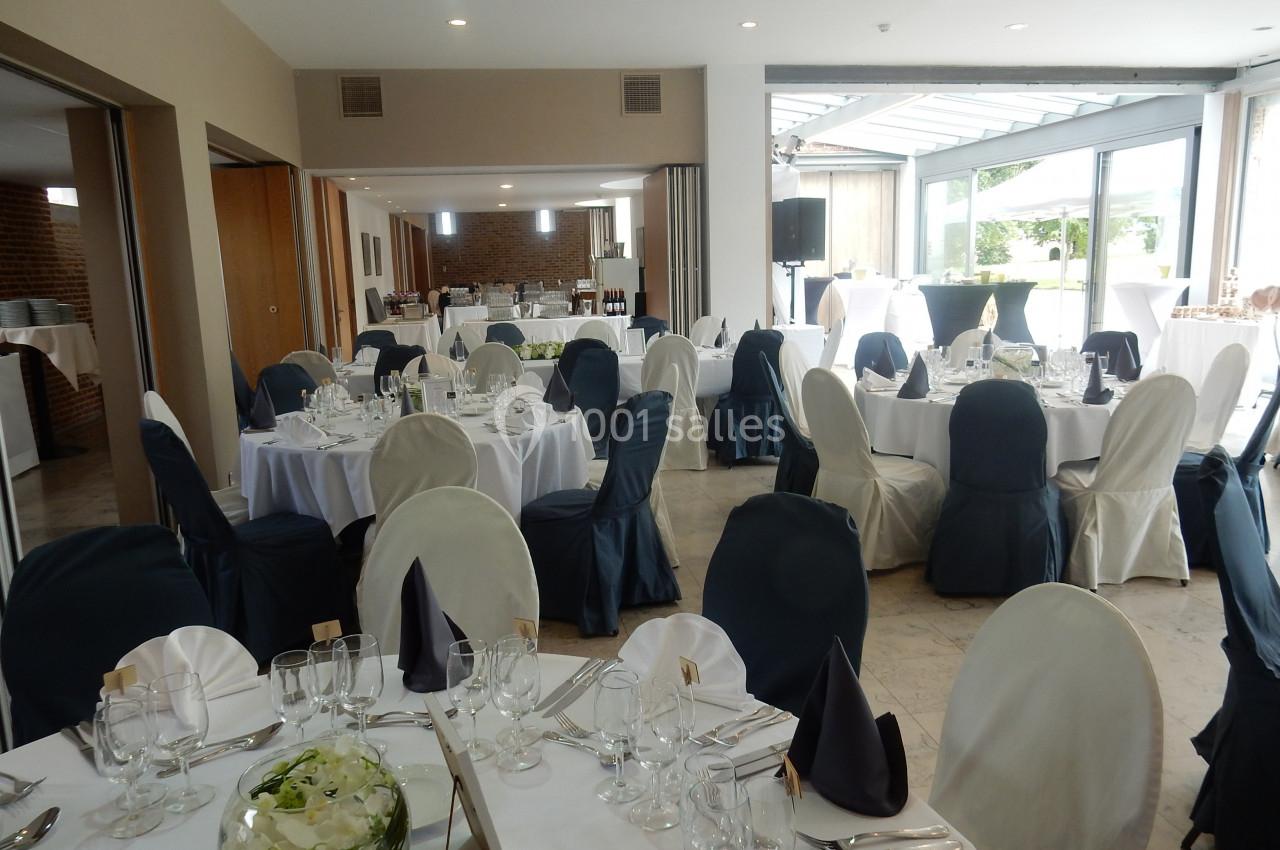 Location salle Donceel (Liège) - Château de Limont #2 Salle de réception lumineuse avec tables rondes dressées, nappes blanches et chaises couvertes de housses noires et blanches.