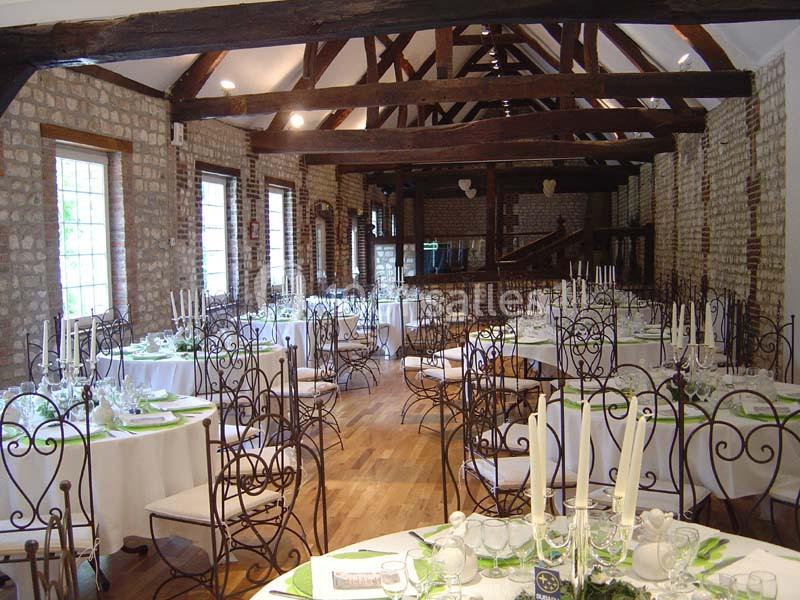 Salle de réception avec poutres apparentes, tables rondes dressées, chaises en fer forgé et lumière naturelle.