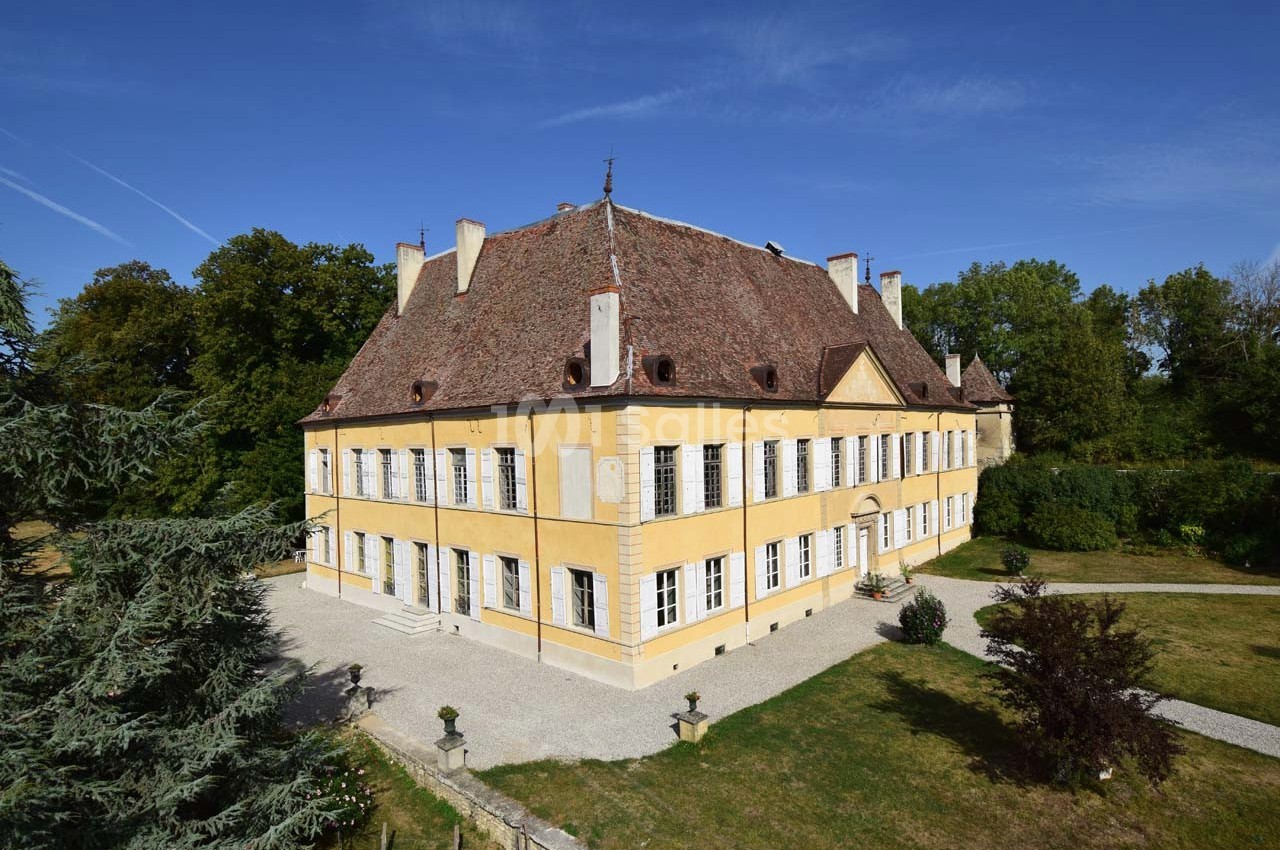 Façade d'un grand bâtiment historique jaune avec toit en tuiles, entouré de jardins et d'arbres sous un ciel bleu.