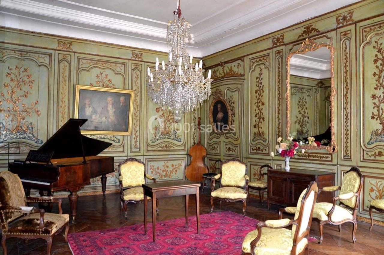 Salon ancien avec boiseries peintes, lustre en cristal, piano à queue, meubles d'époque et tapis rouge au sol.