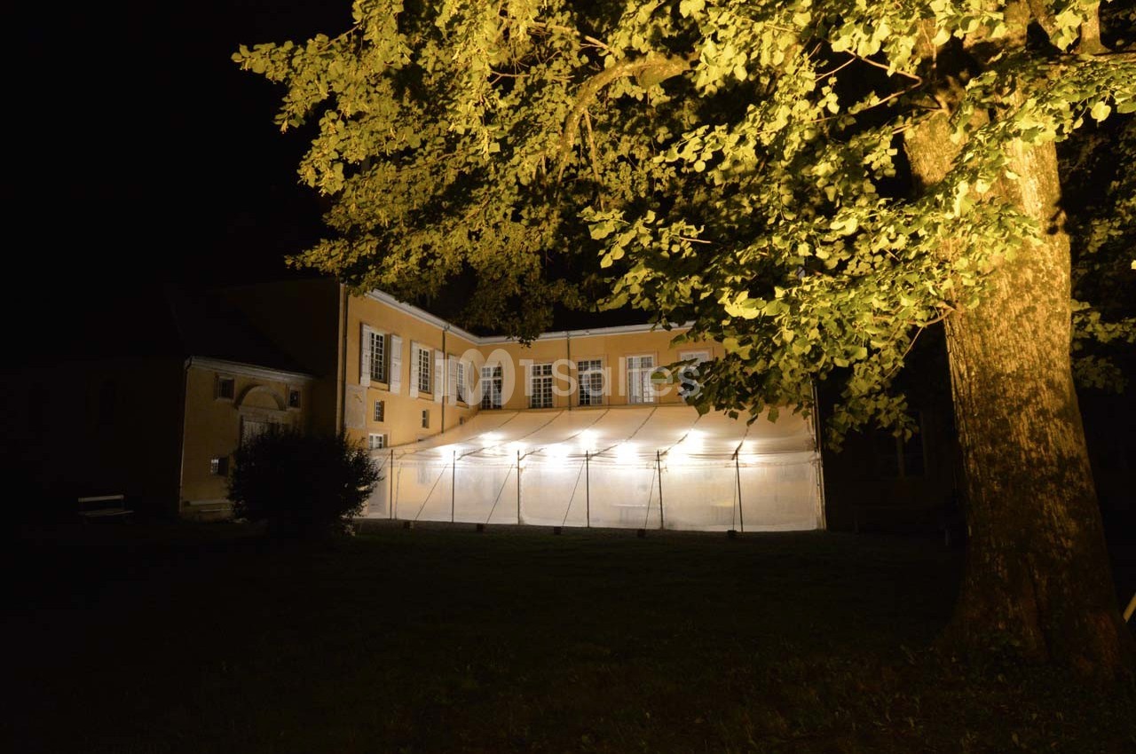 Façade d'un bâtiment éclairé la nuit, avec une tente lumineuse installée dans le jardin sous un grand arbre.