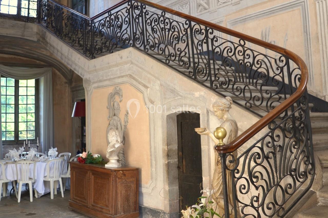 Escalier ancien en pierre avec rampe en fer forgé, statue décorative et table dressée dans un coin lumineux.