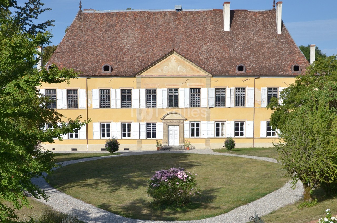Façade d'un bâtiment historique à deux étages avec un toit en pente, entouré d'un jardin et d'allées gravillonnées.
