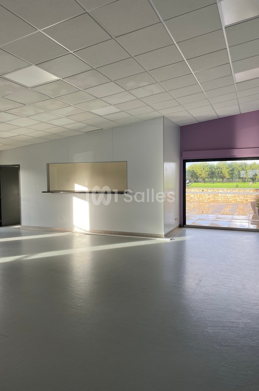 Salle vide avec sol gris, murs blancs, tableau mural, grande fenêtre donnant sur une terrasse et un espace vert.