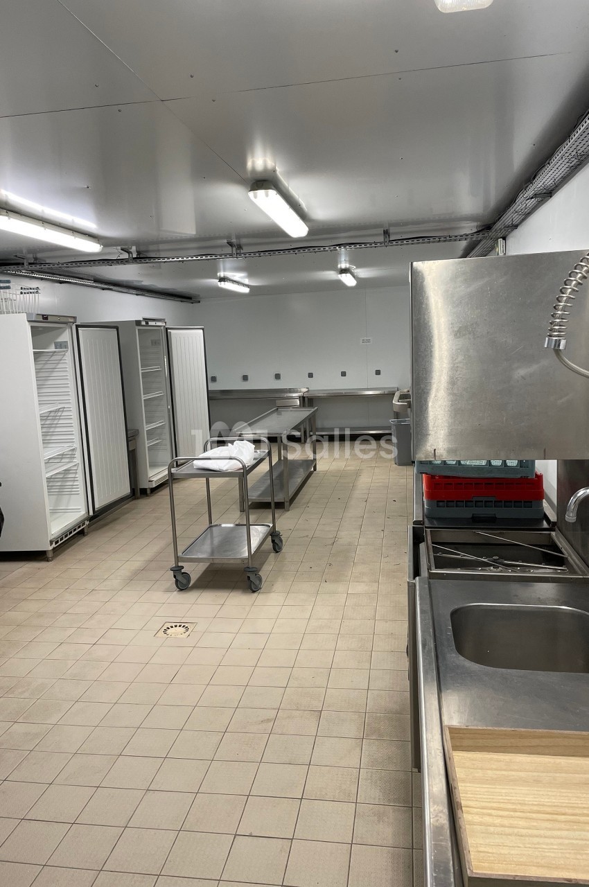 Cuisine professionnelle avec équipements en inox, réfrigérateurs ouverts, plans de travail et sol carrelé beige.