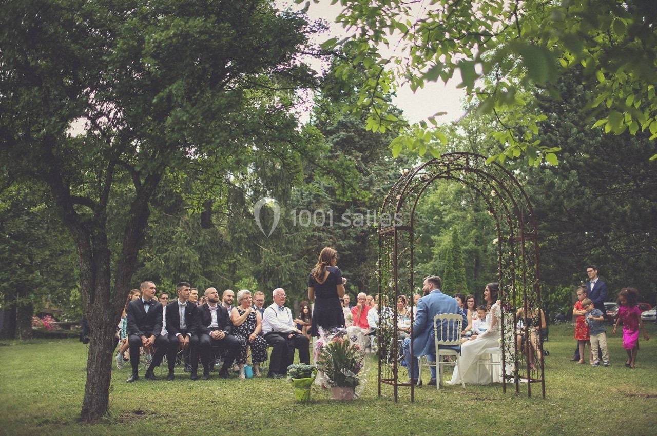 Cérémonie de mariage en plein air avec des invités assis, un couple marié et une officiante sous une arche fleurie.