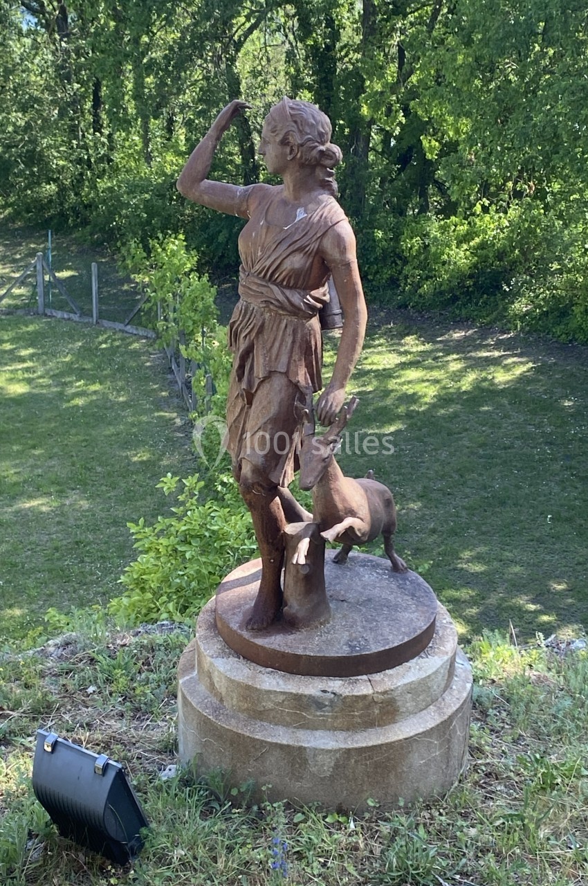 Statue en bronze représentant une femme debout avec un cerf, située dans un parc verdoyant.