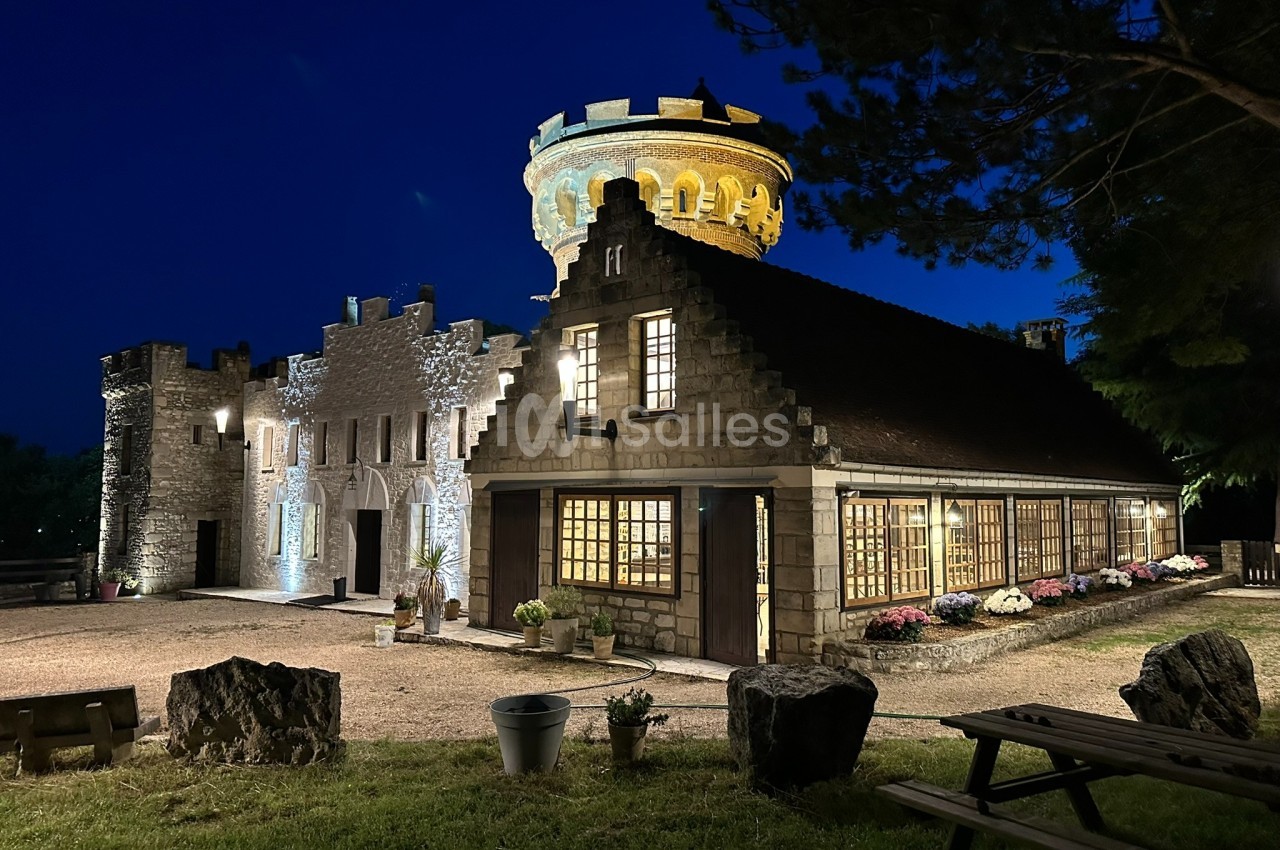 Bâtiment en pierre éclairé de nuit, avec une tour illuminée et des fenêtres vitrées, entouré d'arbres et de mobilier…