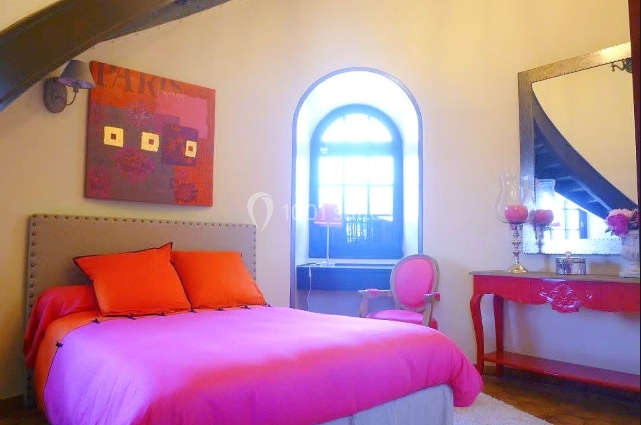 Chambre lumineuse avec lit rose, fauteuil assorti, bureau rouge et grande fenêtre voûtée laissant entrer la lumière.