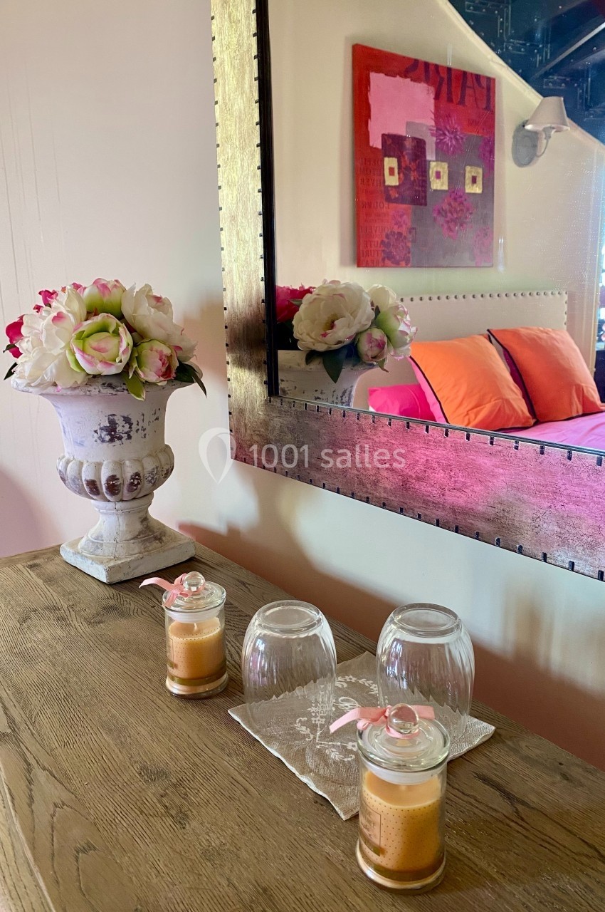Vase de fleurs, bougies et verres posés sur une table en bois devant un miroir reflétant une chambre colorée.