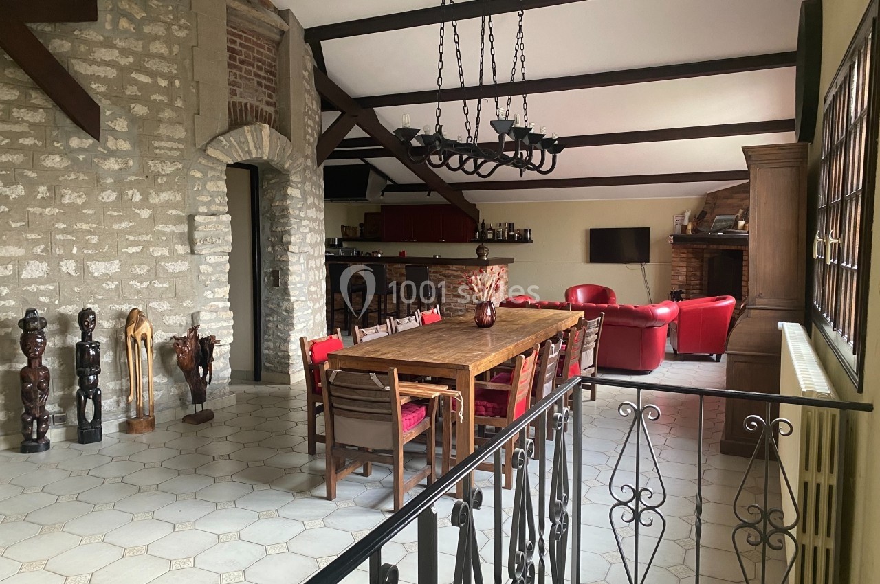 Salle à manger spacieuse avec table en bois, chaises rouges, murs en pierre, poutres apparentes et salon en arrière-plan.