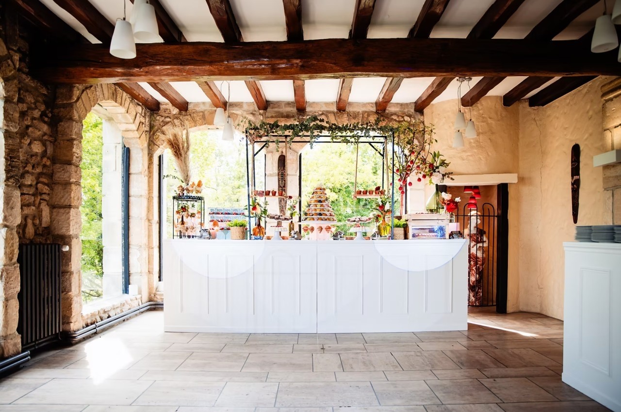 Comptoir de réception décoré avec des desserts et des fleurs, dans une salle lumineuse aux murs en pierre et poutres…