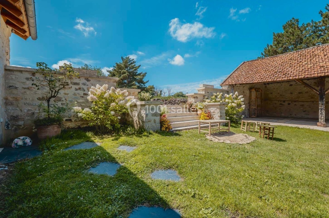 Cour extérieure avec pelouse, fleurs, escaliers en pierre, chaises en bois et bâtiments en pierre sous un ciel bleu.