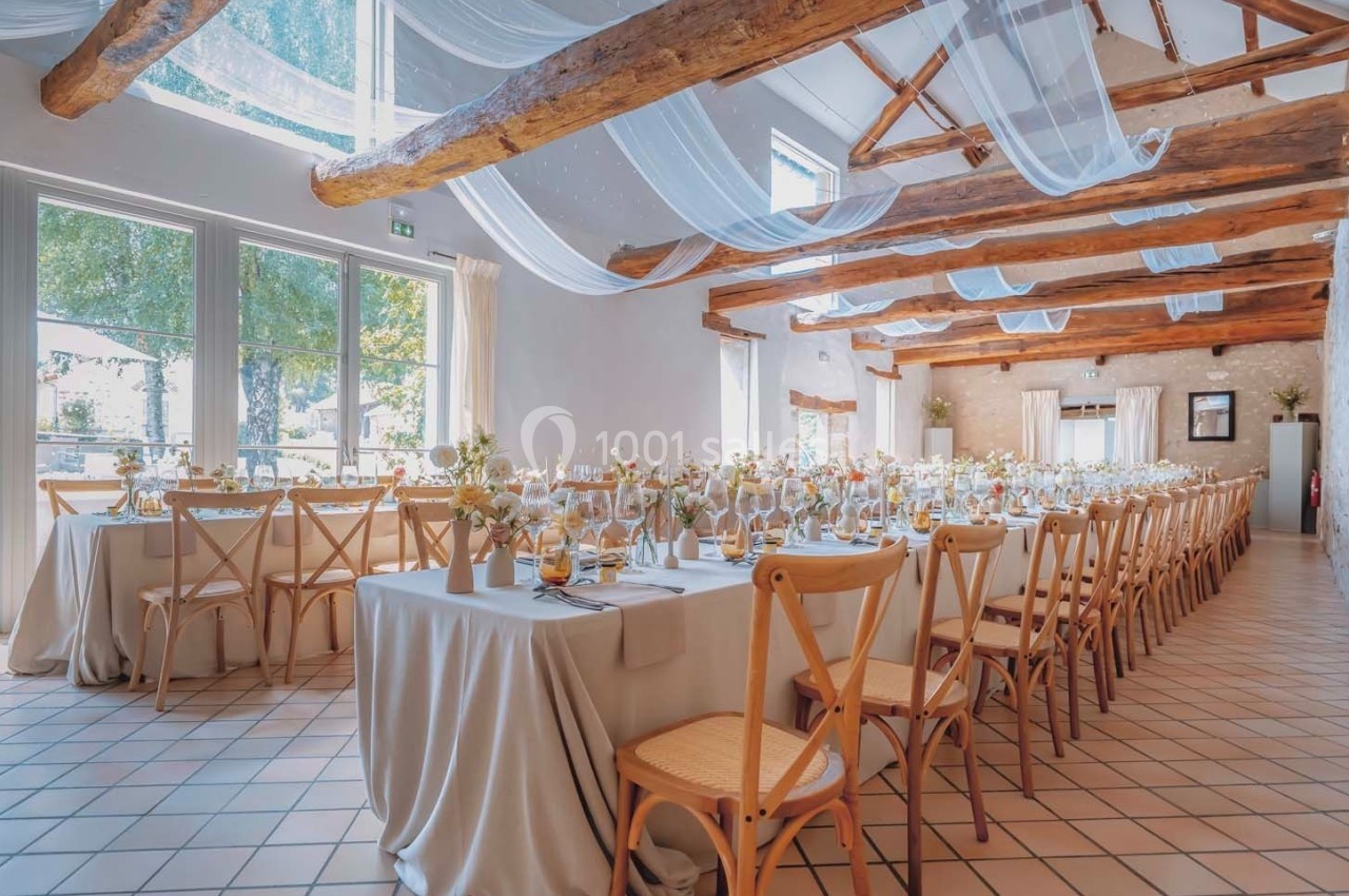 Salle lumineuse décorée pour un événement, avec tables dressées, chaises en bois et voilages suspendus au plafond.