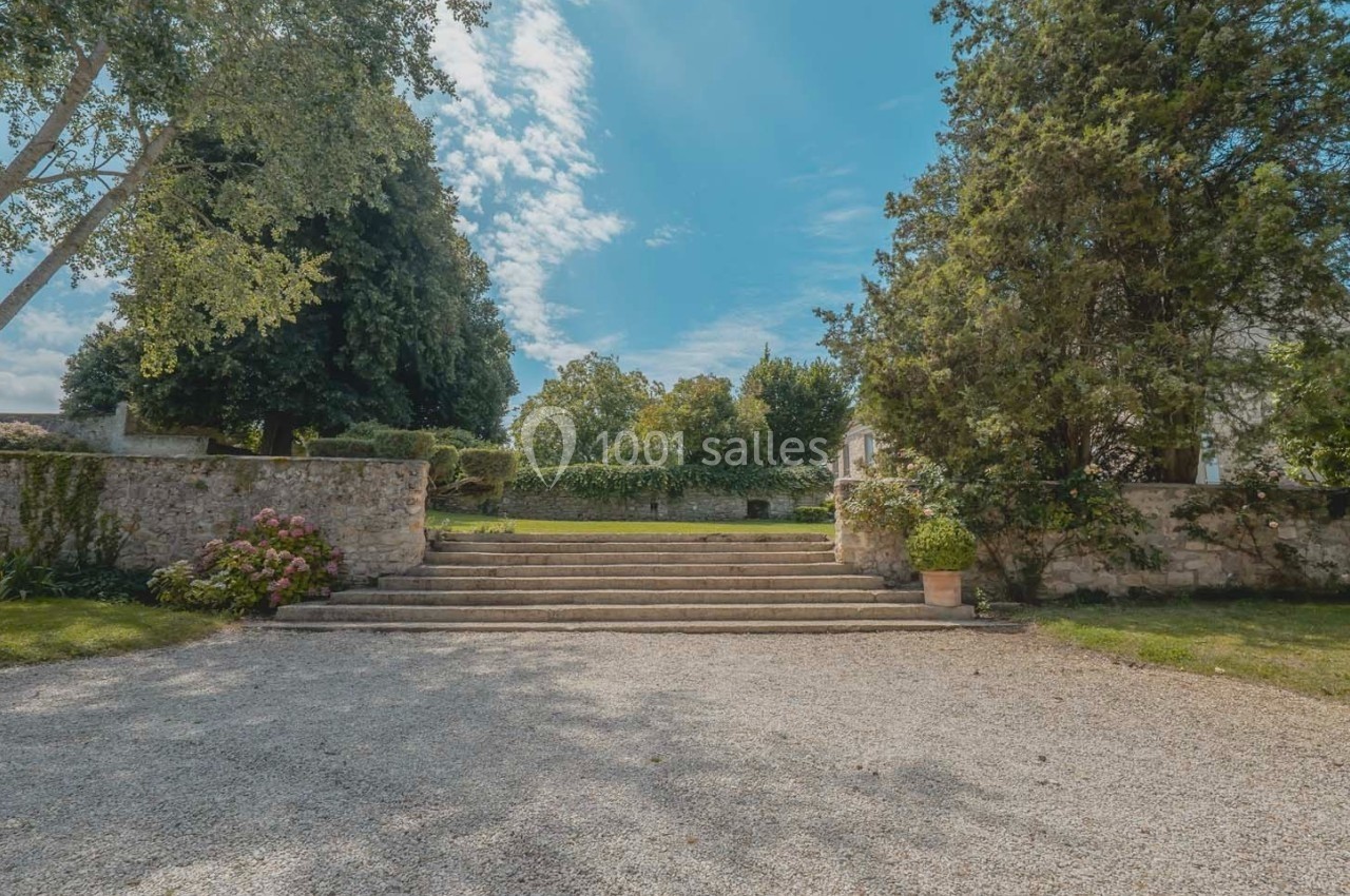 Escalier en pierre entouré de végétation, menant à un jardin avec ciel bleu et quelques nuages en arrière-plan.