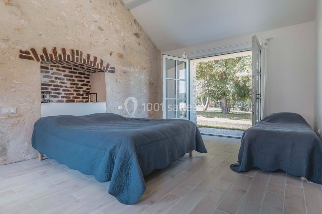 Chambre lumineuse avec murs en pierre, deux lits recouverts de couettes bleues et porte-fenêtre donnant sur un jardin.