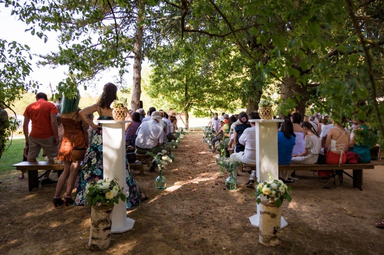 Invités assis sur des bancs en plein air lors d'une cérémonie, entourés d'arbres et de décorations florales.