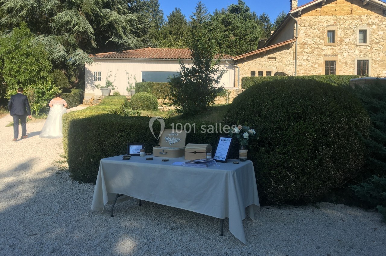 Table avec urne et décorations de mariage devant un bâtiment en pierre, avec des mariés s'éloignant dans un jardin.