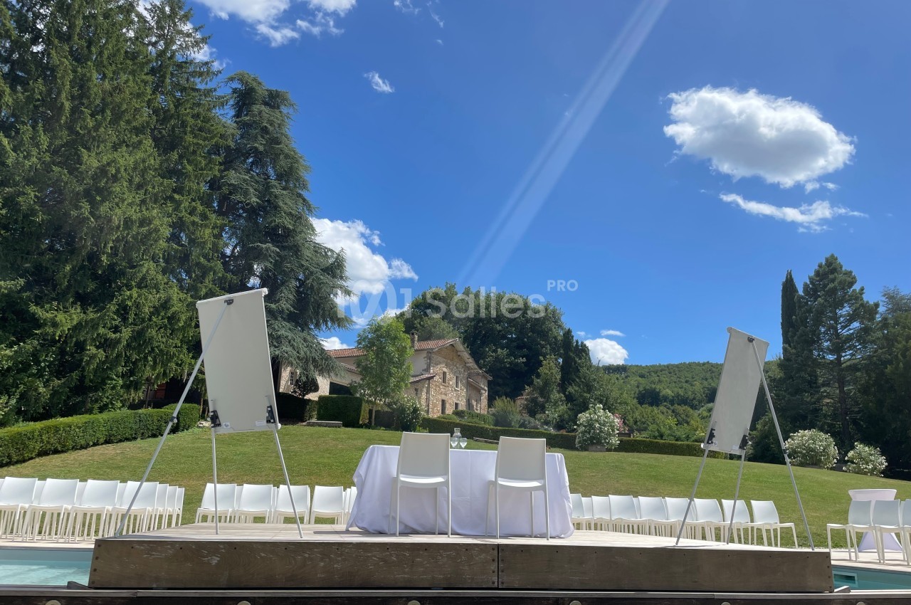 Estrade en plein air avec chaises blanches, tableaux blancs et table, entourée de verdure et d'un ciel dégagé.