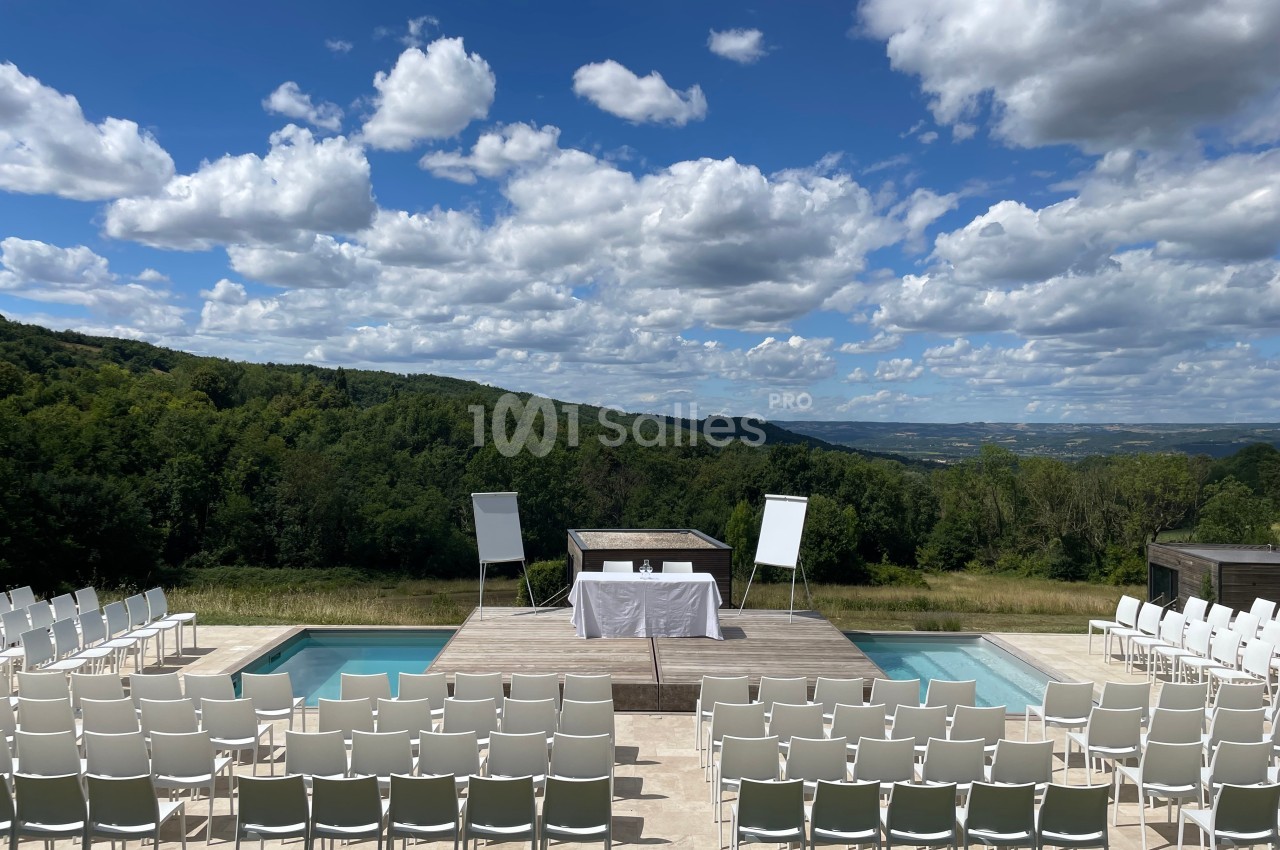 Chaises blanches disposées autour d'une estrade en plein air avec vue sur des collines et un ciel partiellement nuageux.