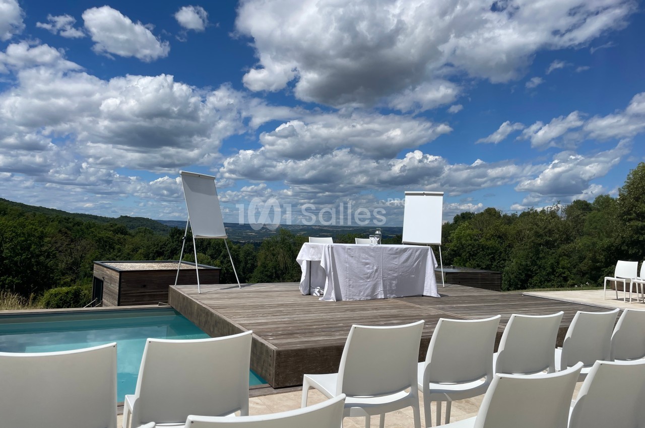 Plateforme en bois avec chevalets et table dressée, entourée de chaises blanches, vue sur un paysage verdoyant.