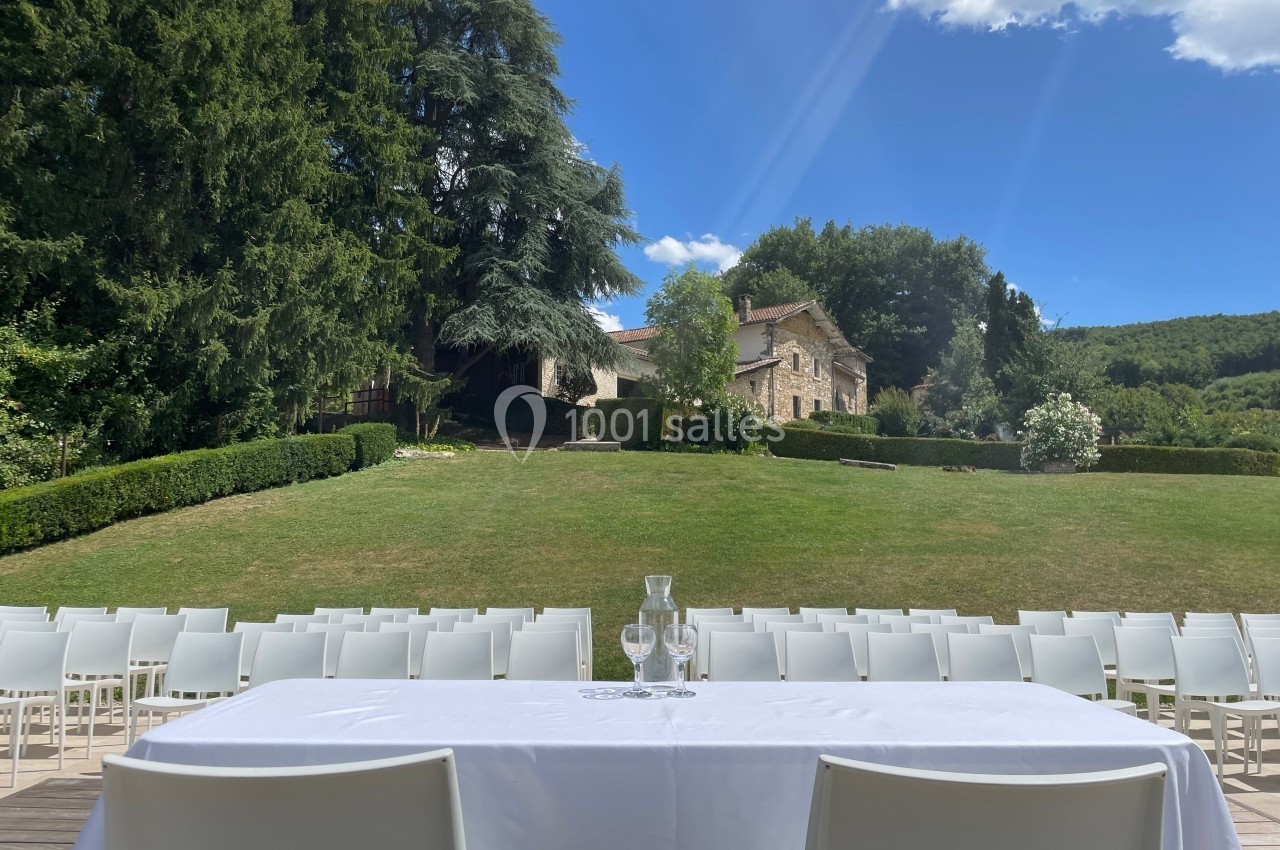 Table dressée en extérieur avec chaises alignées, devant un grand jardin verdoyant et une maison en pierre.