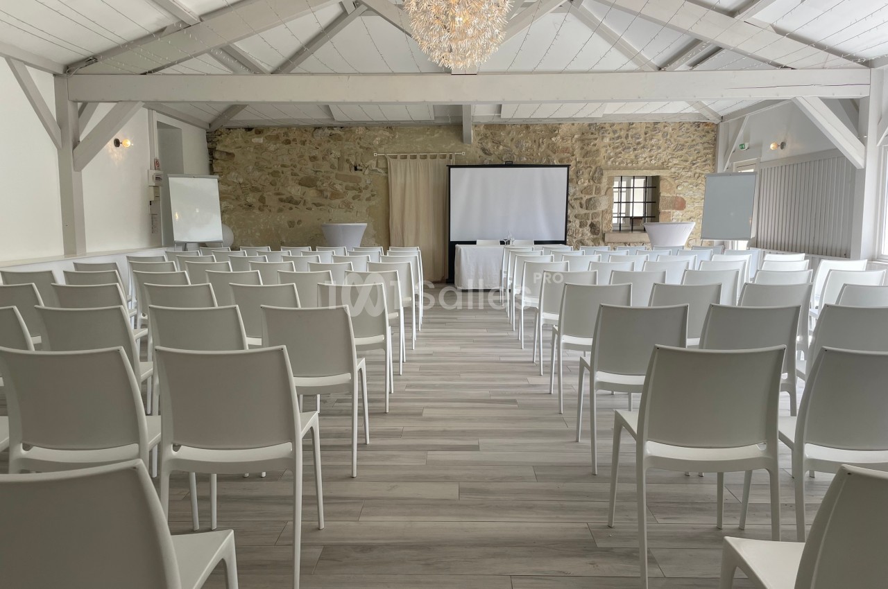 Salle de conférence lumineuse avec des chaises blanches alignées, un écran de projection et des murs en pierre apparente.