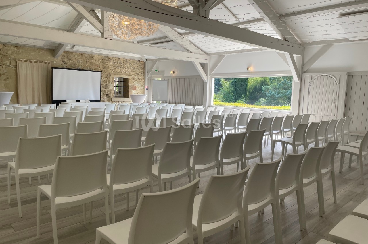 Salle lumineuse avec des rangées de chaises blanches alignées, un écran de projection et une grande baie vitrée.