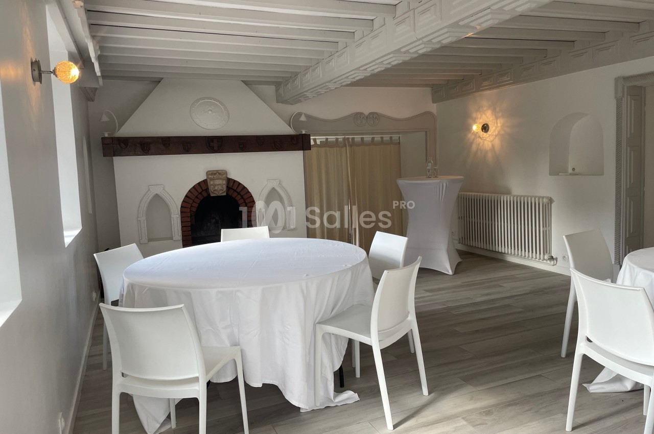 Salle lumineuse avec tables rondes nappées de blanc, chaises modernes et cheminée décorative en arrière-plan.