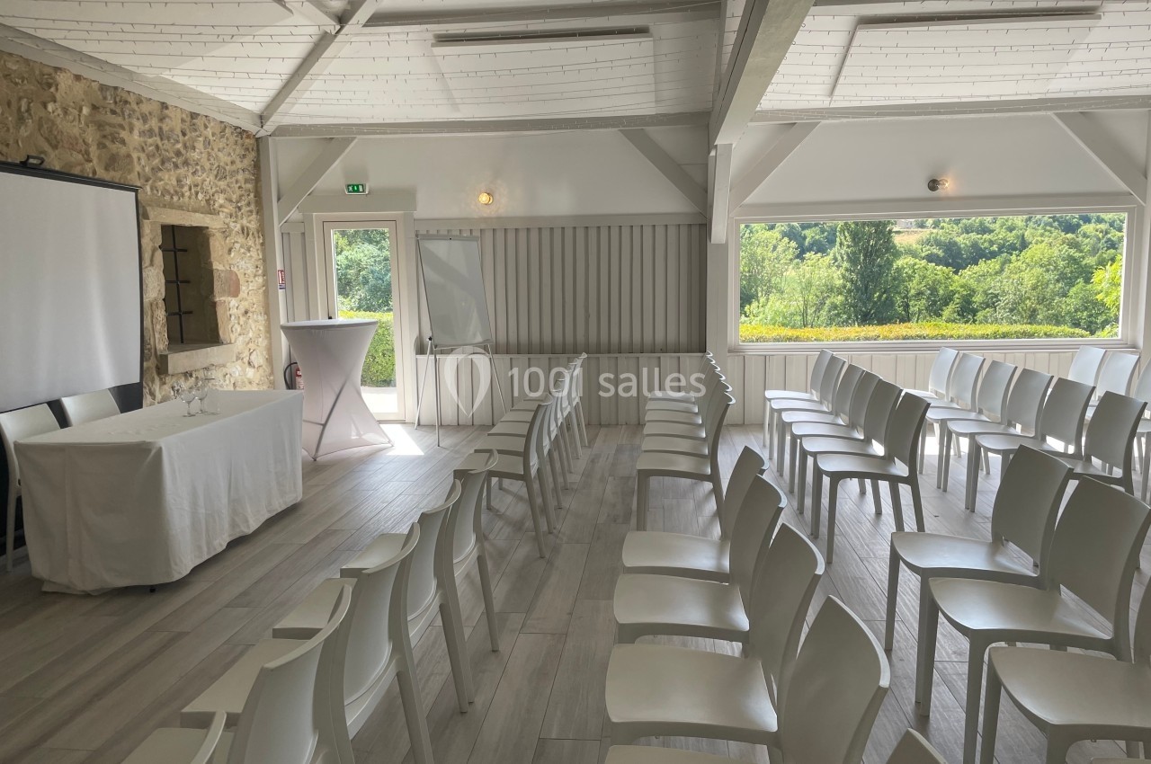 Salle lumineuse avec chaises blanches alignées, écran de projection et vue sur un paysage verdoyant à travers une grande…