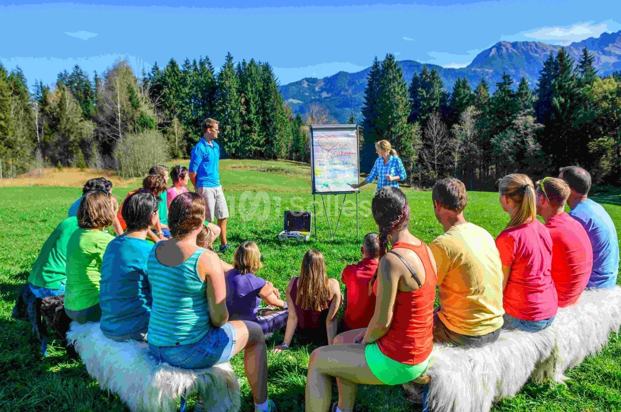 Un groupe de personnes assises en plein air écoute une présentation sur un tableau devant un paysage montagneux.