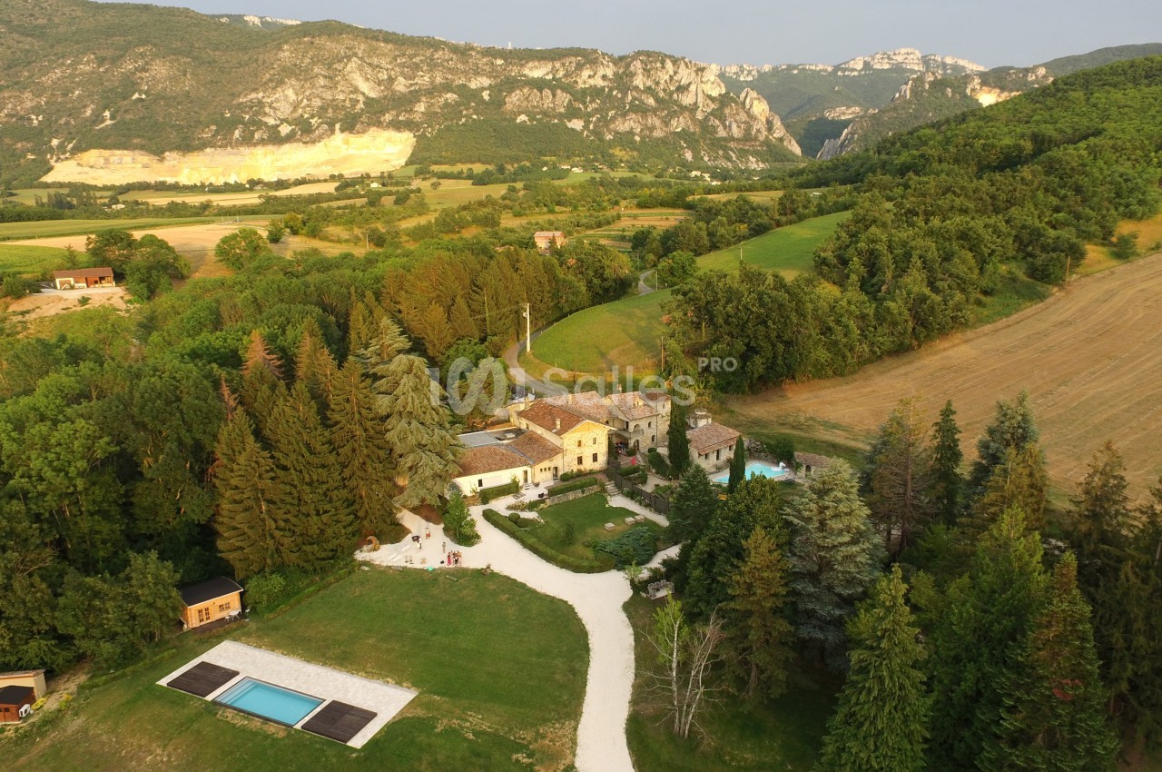 Vue aérienne d'un domaine rural avec bâtiments, piscine, arbres et collines environnantes sous un ciel dégagé.
