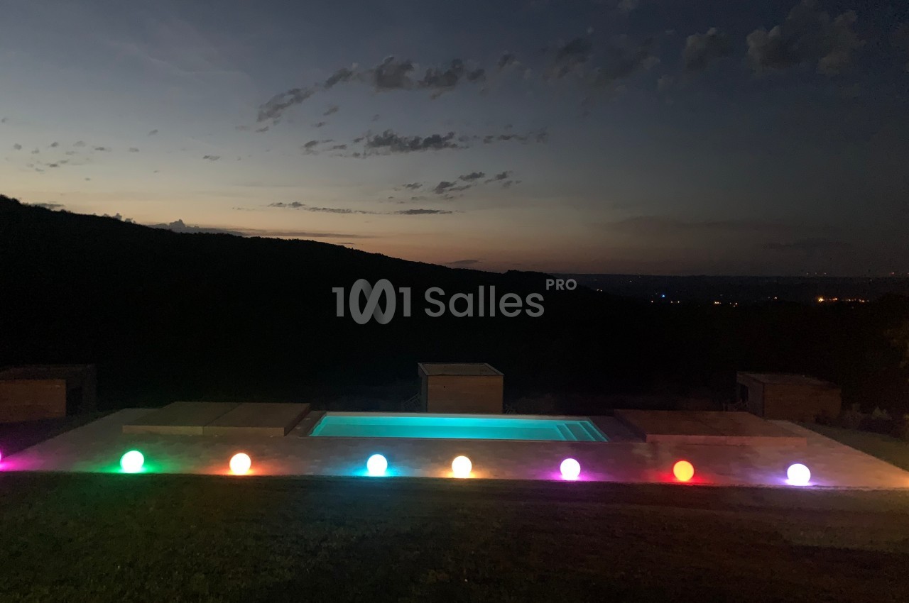 Piscine éclairée par des sphères lumineuses colorées, avec un paysage sombre et un ciel crépusculaire en arrière-plan.