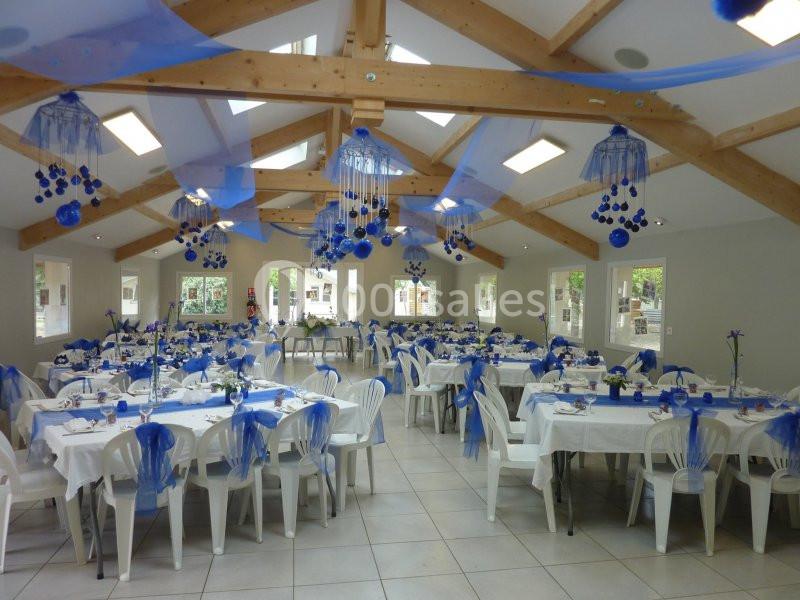 Salle de réception décorée en bleu avec des tables dressées, des chaises blanches et des guirlandes suspendues.