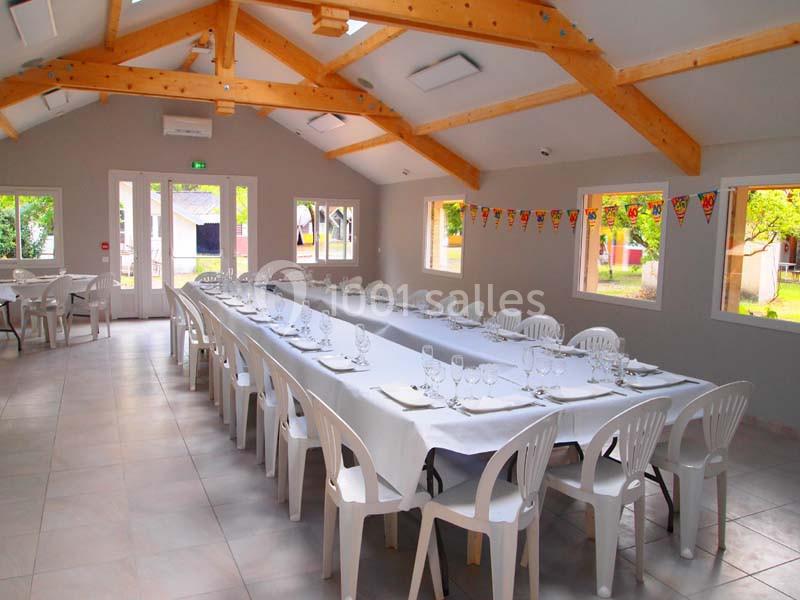 Salle lumineuse avec de grandes tables dressées, chaises blanches et guirlandes colorées accrochées aux poutres en bois.