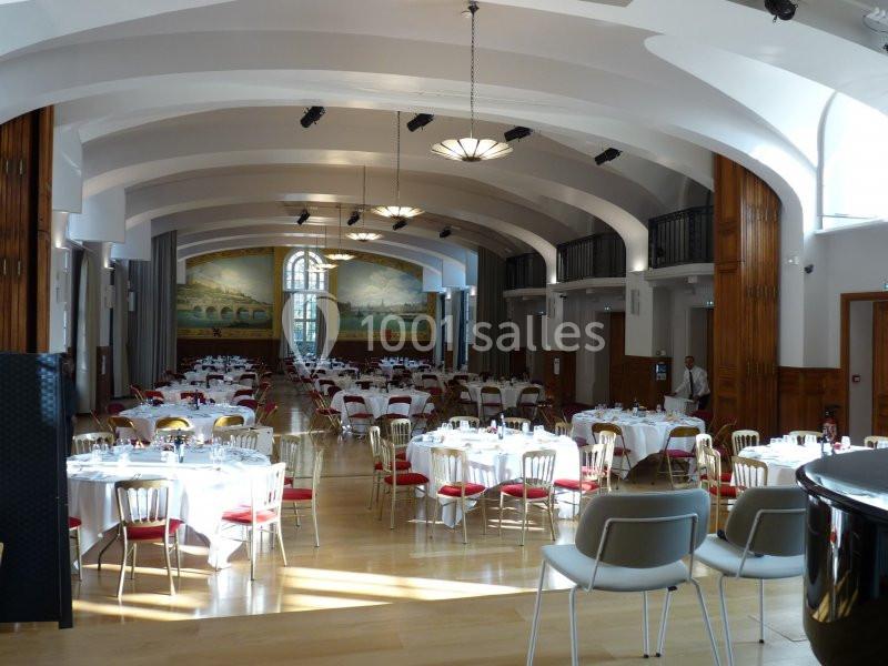 Salle de réception lumineuse avec des tables rondes dressées, chaises blanches et rouges, et une fresque murale au fond.