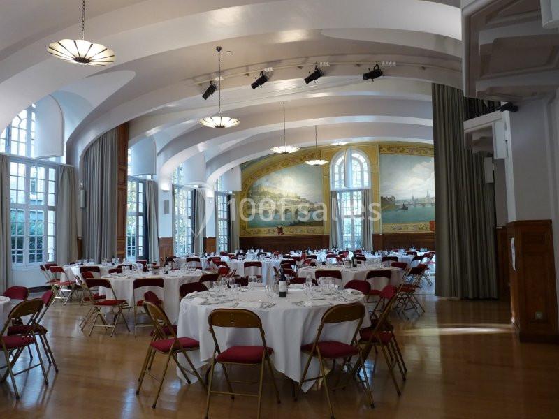 Salle de réception lumineuse avec tables rondes dressées, chaises rouges et fresque murale représentant un paysage.